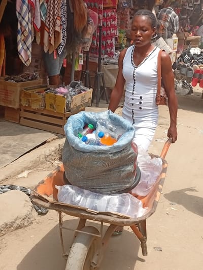 An adult female, appearing to be of African descent, is depicted outdoors in Fegge, Nigeria, actively pushing an orange-colored, single-wheeled wheelbarrow on an unpaved dirt surface. The woman has short, braided hair and wears a sleeveless white dress with black trim around the V-neck and black decorative buttons down the front. A light brown shoulder bag hangs from her left shoulder. The wheelbarrow's tray contains a large, grey mesh bag with a blue plastic lining filled with various plastic bottles of beverages, including water and orange-colored liquid. Several clear plastic bags containing other packaged items are also present in the wheelbarrow. In the background, to the left, market stalls display hanging scarves, wigs, and stacked cardboard boxes filled with merchandise. To the right, another stall exhibits numerous pairs of sandals on racks. The scene is brightly lit by daylight, casting shadows.