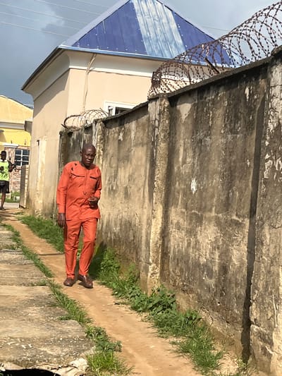 In Abuja, Nigeria, an older Black man walks along a dirt path beside a high concrete wall topped with barbed wire. He is wearing an orange two-piece outfit with black trim. A younger Black person wearing a neon green shirt and dark shorts is visible in the background to the left. The scene is outdoors, with a cloudy sky and a building with a blue metal roof visible behind the wall. The wall itself is weathered and stained, with patches of white paint or graffiti. The path is lined with sparse green vegetation.