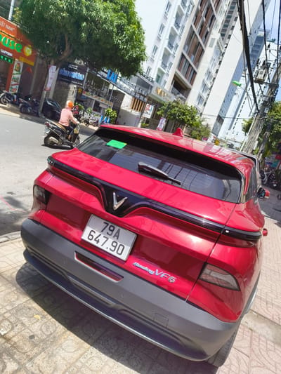 A red VinFast VF8 electric SUV is parked on a sidewalk next to a street in Nha Trang, Vietnam. The car has a license plate reading "79A 647.90". In the background, there are buildings, trees, and other vehicles. A person is riding a motorcycle in the street, and several parked motorcycles are visible. The environment appears to be a sunny day in a city.