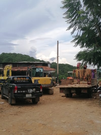This image depicts an outdoor scene in what appears to be a yard or depot, likely in Da Lat, Vietnam, given the text on the vehicles. The primary subjects are several construction vehicles, including two yellow excavators and a black pickup truck. A flatbed truck loaded with materials, possibly pipes or scaffolding, is also prominent. A motorcycle is parked to the right.
The setting is a dusty, unpaved area with a building featuring a corrugated metal roof in the background. Lush green hills covered in trees form the backdrop under a cloudy sky. The time of day appears to be overcast, possibly late morning or afternoon, with the weather seeming humid and potentially leading to rain.
The vehicles suggest an active work environment, possibly related to construction, repair, or landscaping. The black pickup truck has visible text on its tailgate and rear window. On the tailgate, text reads "XE ĐI CÂY!", which translates to "Tree Going Vehicle!" or "Vehicle for Trees!". It also displays a license plate "77A 478.60". On the rear window, there is text that appears to be advertising or service information for "Phúc Thủy Lực" (Phuc Thuy Hydraulic), offering "SỬA CHỮA XE CƠ GIỚI" (Repair of Mechanical Vehicles) with phone numbers and a location mentioning "ĐÀ LẠT - LÂM ĐỒNG" (Da Lat - Lam Dong). One of the excavators has a "VOLVO" logo and an "EX35" model designation visible.
The overall impression is of a functional, working yard preparing for or engaged in heavy-duty tasks, possibly involving tree removal or other land-based operations.