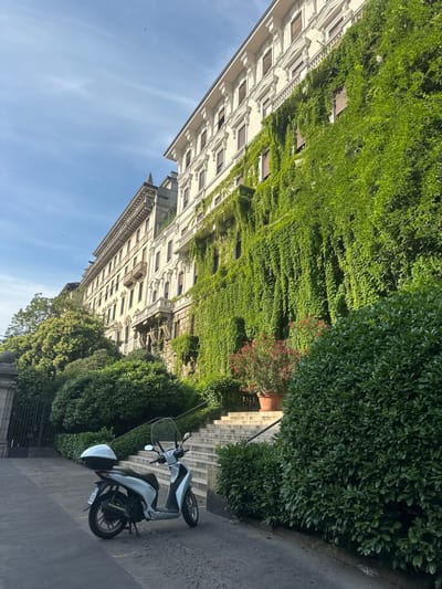 This photograph depicts a grand urban scene in central Milan, Italy, likely along or near Via Vittorio Veneto or Via Manzoni. The image is dominated by a multi-story building featuring classical Italian architecture, including ornate cornices, decorative window frames, and balconies with intricate railings. A significant portion of the building's right facade is enveloped in a dense curtain of vibrant green ivy, which cascades downwards and partially obscures numerous windows fitted with dark shutters.
To the left of the heavily ivy-covered section, the building's cream-colored facade is more visible, showcasing additional windows and a prominent central balcony. Below the building, a wide, multi-tiered concrete or stone staircase ascends towards the right, partially bordered by a manicured dark green hedge. A black metal handrail is discernible alongside the stairs. At the base of the staircase, a terracotta pot contains a bush with small red flowers.
In the lower left quadrant of the image, a light blue and white Honda SH scooter is parked on a dark paved surface, oriented slightly uphill towards the right. The scooter is equipped with a clear windscreen and a white top box attached to its rear rack. Further to the left, a large, dark metal gate is partially visible, situated behind dense green foliage and trees.
The sky is a light, clear blue with faint, wispy white cloud formations. A small dark bird is visible in flight in the upper left portion of the sky. The scene is brightly illuminated by daylight, creating shadows that indicate either morning or late afternoon sunlight. No individuals are present in the frame.