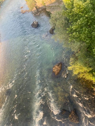 The image is an aerial view of a river flowing through a natural environment. The river has clear, greenish-blue water with visible currents and some white foam suggesting moderate flow. Several large rocks are submerged in the river, breaking the surface in places. The riverbanks are lined with lush green trees and vegetation, with a rocky, sunlit cliff face visible on the upper left side. The lighting suggests it is either early morning or late afternoon, with sunlight casting warm tones on the vegetation and cliff. There are no people or man-made structures immediately visible. This scene is consistent with the Moraca River in Podgorica, Montenegro, particularly in areas where it flows through more natural and less urbanized sections, characterized by rocky outcrops and dense riverside foliage.