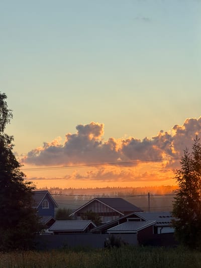 The image depicts a rural or suburban landscape at sunset. The sky is a gradient of soft blues, yellows, and oranges, with dramatic, illuminated clouds. In the foreground, the tops of several houses with gray roofs are visible, nestled among trees. A dark fence runs horizontally across the middle ground, separating the houses from a field of tall grass in the foreground. Electrical wires are strung across the scene, spanning between utility poles. The overall impression is one of a tranquil evening in a residential area. This scene is not located in the city of Mars, Russia, as Mars is a planet, not a city in Russia.