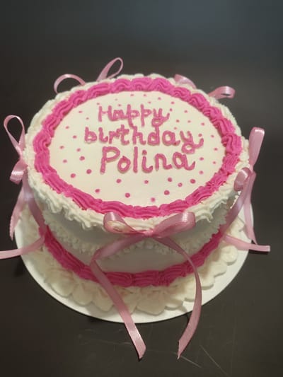 A two-tiered white cake with pink frosting decorations and pink ribbons tied around it sits on a white cake board. The top tier of the cake has "Happy birthday Polina" written on it in pink frosting. The cake is centrally framed and shot from a slightly elevated angle. The background is a dark, textured surface that appears to be a table. There are no people visible in the image. The scene is indoors, with no discernible architectural features indicating a specific location. There is no information available to determine if this image is located in the city of Myadzyel, Belarus.
