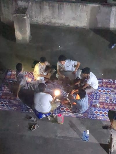Six men are sitting on a colorful patterned blanket spread out on a concrete surface, illuminated by a lamp. The men are all wearing casual clothing and appear to be conversing. Two empty plastic cups and a pink water bottle are visible on the blanket. Two bottles of water are on the concrete floor, to the right of the blanket. The location is a rooftop in Dar es Salaam, Tanzania. The concrete surface appears to be a rooftop or a balcony. The setting sun casts a warm glow on the scene.