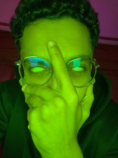 A close-up, portrait-oriented image depicts an individual from the chest up, heavily saturated by a bright neon green light. The person has dark, short, curly hair and wears round, metal-rimmed eyeglasses. Their eyes, visible through the lenses, appear luminescent green, reflecting a blue and white pattern indicative of a sky or clouds. The right hand (viewer's left) is raised, with the index finger pressed vertically against the forehead, covering the bridge of the nose. The left hand (viewer's right) rests against the left side of the face, fingers slightly curled, partially obscuring the lower cheek and jawline. The individual wears a dark, hooded garment, visible on the shoulders and upper chest. The background is indistinct, displaying a blurred magenta-purple hue, consistent with an indoor environment illuminated by colored lighting. The scene is located in the city of Fez, Morocco.