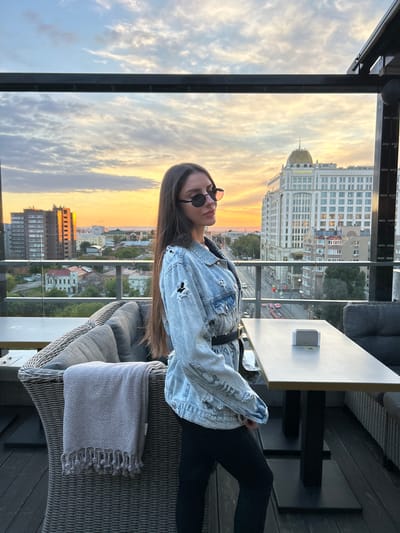 A woman is sitting on a wicker chair on a balcony overlooking a city at sunset. She is wearing sunglasses and a denim jacket. The sky is filled with dramatic clouds and the warm colors of sunset. In the background, there are buildings and a street with cars. The overall mood is serene and relaxed.
The woman has long, dark hair and is wearing a light-wash denim jacket with distressed details. She has on dark sunglasses and appears to be looking towards the sunset. Her posture is relaxed as she leans back in the chair.
The setting is an outdoor balcony or patio with a view of a city. There is a table to her right with a small white object on it, possibly a tissue dispenser or ashtray. A grey blanket is folded on the chair next to her. The architecture in the background includes a large, ornate building with a golden dome, reminiscent of historical European architecture. Other buildings are more modern, with visible windows. The sky shows the vibrant colors of sunset, with streaks of orange, yellow, and pink. The time of day is clearly sunset.
There is some text visible on the golden-domed building. It appears to be in Cyrillic script and reads "КОШЕЛЕВ". This likely refers to the name of the building or a company associated with it. The location is identified as Novokuybyshevsk, Russia.