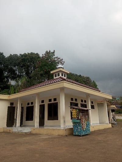 The image displays a single-story building with pale yellow walls and a reddish-brown tiled roof located in Kandapola, Sri Lanka. A small, multi-tiered tower featuring a silver-colored dome and finial crowns the roof. The structure has multiple rectangular, dark-framed windows and two visible dark brown doors. A covered overhang extends around parts of the building, supported by pillars. In front of the building, to the right, a patterned blue vending cart containing various packaged snack items is positioned. The foreground consists of light brown, unpaved ground. Behind the building, dense green trees are visible, and distant residential structures can be seen on a hillside to the right. The sky is uniformly overcast and grey. No people are present in the scene.