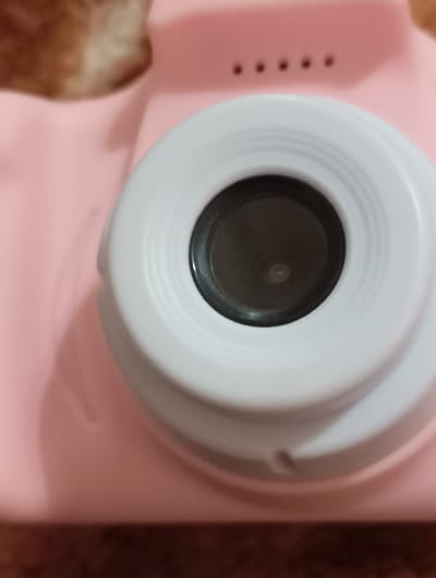 The image depicts a close-up, high-angle view of a children's digital camera. The camera body is uniformly light pink. A prominent white, circular lens housing is centrally located on the camera's front, containing a dark, recessed lens element. Above the lens housing, four small, dark, circular features are arranged horizontally on the pink camera body, possibly indicating lights or microphone apertures. The camera rests on an out-of-focus, flat, reddish-brown surface. A blurry, light brown object is partially visible in the upper left portion of the frame. No individuals are present. The scene is located in the city of Kurganinsk, Russia.
