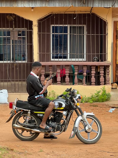 A young man is sitting on a black motorcycle, holding a phone. He is wearing a black t-shirt with white and red trim and black shorts. The motorcycle has a silver exhaust pipe and a silver front wheel with black spokes.
The man is positioned in front of a building with a cream-colored facade. The building has barred windows with dark panes. The ground is a mix of dirt and patchy green grass. There are also some plants growing near the building.
The image appears to have been taken during the daytime, likely late morning or afternoon, given the bright lighting. The weather seems to be fair, with no visible rain or clouds.
The setting is likely a residential area in Nkpor, Nigeria, suggested by the building's architecture and the dirt road. There is no clear indication of emotion from the man, as he is focused on his phone.
Visible text includes "SUPER" on the motorcycle's front wheel, and some unreadable text on the side of the motorcycle.