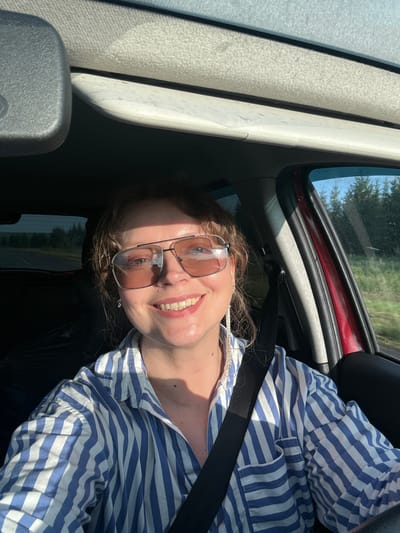 A woman is driving a car. She is wearing sunglasses and a blue and white striped shirt. She has a seatbelt on. The sun is shining through the windshield. The car appears to be on a road in a rural area, possibly in Verkhnie Iziverki, Russia, given the trees and the natural landscape visible outside the windows. The image is a selfie, taken from inside the car.