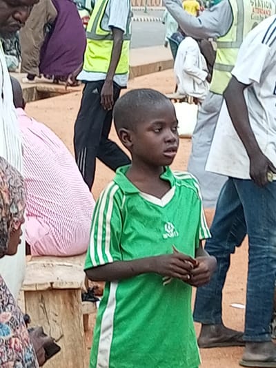 This is a medium shot of a group of people in what appears to be a market or busy street area in Gombe, Nigeria. In the foreground, a young boy is looking to his left, wearing a green t-shirt with white stripes on the sleeves and a white trim around the neck. His hands are clasped in front of him. To his left, a person is wearing a pink and white striped garment. Behind the boy, a person wearing a bright yellow vest over a white shirt and black pants is visible. Further in the background, other individuals are present, some wearing traditional Nigerian attire, and a building with signage indicating "STATION" is visible. The ground is reddish-brown earth, suggesting an outdoor environment.