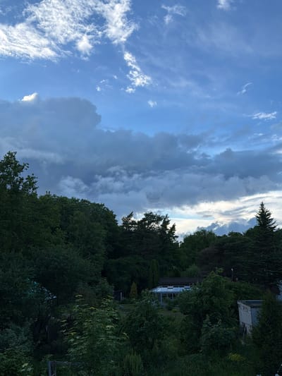 The image shows a view from a high vantage point overlooking a densely wooded area with some structures visible. The sky is filled with a mix of dark, dramatic clouds and lighter, wispy clouds, suggesting an impending storm or recent rainfall. The trees are lush and green, indicating a temperate climate. In the middle ground, a modern house with a conservatory or sunroom is visible, nestled amongst the trees. To the right, a smaller, utilitarian building, possibly a shed or outbuilding, with a light-colored, corrugated roof is present. The overall scene is one of a rural or semi-rural residential area with significant natural vegetation. This image does not provide any specific visual cues that would allow for an identification of the location within the city of Tallinn, Estonia.
