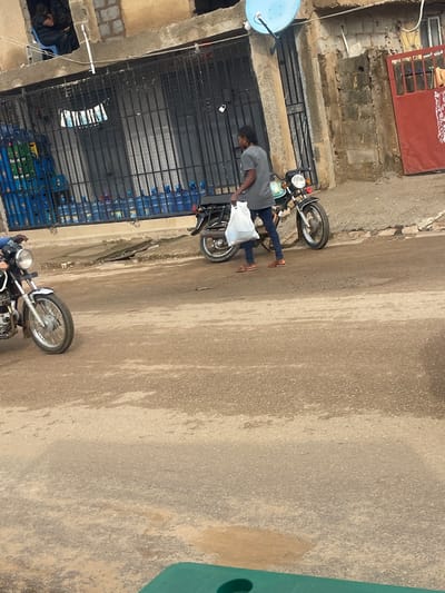 A person is walking on a paved street in front of a building with barred windows. The person is carrying a white plastic bag and is wearing a grey t-shirt and blue jeans. A motorcycle is parked next to the person. The building appears to be a shop or residence, with stacks of blue plastic water bottles visible behind the bars. The street is worn and cracked in some areas. The overall scene suggests an urban environment, possibly in Abuja, Nigeria, given the water bottle branding if any were discernible.