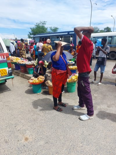This image captures a vibrant, bustling street scene in Dar es-Salaam, Tanzania, under bright daylight with a partly cloudy sky. The perspective is slightly elevated, looking down into the street.
The scene is densely populated with people and activities, suggesting a market area or a busy public transport hub. In the foreground, a man wearing a red polo shirt, purple trousers, and white Crocs stands with one arm raised to his head. Beside him, a woman in a blue top and a red, patterned skirt stands next to several green and red buckets overflowing with packaged snacks, resembling popcorn or puffed rice. She is wearing black leggings and black slide sandals with "DIOR" visible on one.
Further back, numerous other street vendors are visible, some seated, also surrounded by buckets of similar packaged goods. A man in a black t-shirt with "LIVING THAT" and "NEW YORK" visible text, and denim shorts, is seen holding a large blue multi-gallon water bottle.
Several buses are parked or moving in the background, indicating a transport-oriented setting. People are congregated around these buses. At the very bottom edge of the frame, the front of a multi-colored vehicle, possibly a tuk-tuk or Bajaj, is partially visible, with "TRAVEL" or similar text on its side. Street lamps extend into the sky. The ground is paved asphalt. The overall atmosphere is one of lively commerce and daily urban activity.
