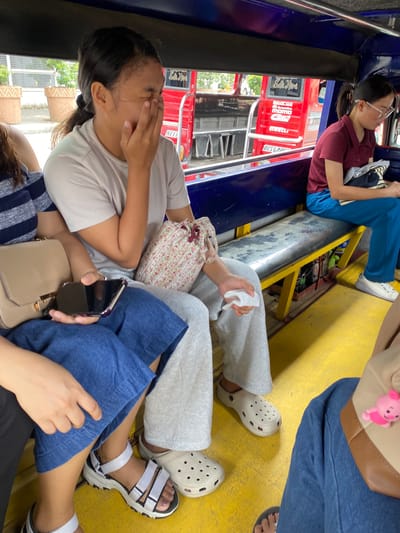 The image shows the interior of a public jeepney in Davao City, Philippines, filled with passengers. In the foreground, a young woman is seated, covering her face with her hand, possibly yawning or in distress. She is wearing a light-colored t-shirt and grey sweatpants, with white Crocs. Beside her, another person is partially visible, wearing a blue denim-like skirt and white sandals. In the middle ground, a woman in a maroon polo shirt and blue pants is seated, wearing glasses and holding a bag. The jeepney's interior is characterized by blue plastic seating and a yellow floor. Outside the jeepney, through the windows, glimpses of a red vehicle with advertisements and planters can be seen. The overall scene depicts a typical commute in Davao City.