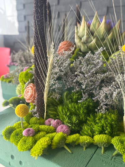 A close-up, vertically oriented shot displays an arrangement of dried and fresh flowers. An artichoke and delicate, wispy seed heads add height. Grayish dried flowers are intertwined with vibrant green, button-like chrysanthemums. A few pink pom-pom flowers and a yellow ball-shaped flower add pops of color. A peach-colored faux rose is present. The flowers are arranged in green floral foam. The context of location in Mexico City is unsupported.