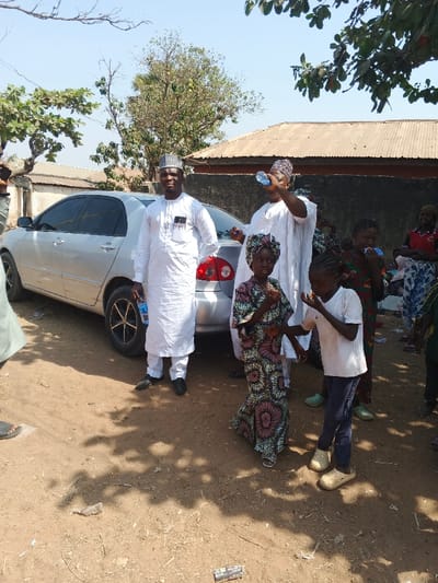 The image displays an outdoor scene in Doma, Nigeria, under clear daylight conditions. A silver four-door sedan, consistent with a Toyota Corolla model, is parked on an unpaved, light brown dirt surface which is partially shaded by an unseen tree.
In the foreground and midground, six individuals are clearly visible, with several more partially seen in the background. To the left of the vehicle, a man wearing a white traditional garment (agbada-style) and a patterned cap stands, holding a clear plastic water bottle in his left hand. His gaze is directed towards the viewer. To the right of the sedan's rear, another man, similarly dressed in a white traditional garment and cap, is actively drinking from a clear plastic water bottle, tilting his head back.
In front of these adults, two children stand in the immediate foreground. A young girl, wearing a patterned dress (featuring circular motifs in brown, pink, and white) and a matching headwrap, holds a small food item to her mouth. Beside her, a young boy in a white t-shirt, dark blue trousers, and light-colored slip-on shoes (crocs) also eats a similar small, irregular food item.
In the background, behind the main group, multiple individuals, predominantly children, are gathered. Some hold clear plastic bottles. They are positioned in front of a low, dark-colored wall and a single-story building with a corrugated metal roof. Another building with a similar roof is visible further left in the background. Sparse trees and shrubs are present in the background, with one tree branch extending into the upper right corner of the frame. The ground shows scattered debris, including a small piece of dark plastic or paper.