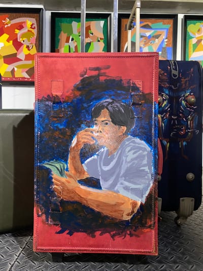 In this image, a red leather-bound book with a painted portrait of a man holding a drink is the central focus. The man has dark hair and is wearing a light-colored shirt. He is depicted with visible brushstrokes, suggesting an artistic rendering. The background of the painting is abstract and colorful, with blues and darker tones. The book appears to be standing upright on a textured metal surface, possibly a floor, with what looks like luggage or suitcases behind it. The suitcases are dark blue and feature some decorative elements, including what could be stylized faces or patterns. In the background, there are framed abstract artworks displayed on a wall, adding to a gallery-like or art exhibition setting. One of the artworks shows geometric shapes in vibrant colors like yellow, orange, and pink. Another artwork features green and grey geometric forms. The lighting suggests an indoor environment, potentially a public space or a personal collection area. The scene is likely located in Davao City, Philippines, based on the context provided, hinting at a cultural or artistic event in the city.