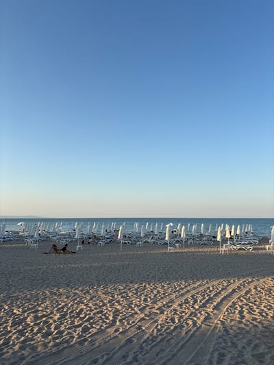 This media file depicts a scenic beach in Sozopol, Bulgaria, likely during the late afternoon or early evening, as indicated by the long shadows cast across the sand. The image is oriented vertically, with the sky occupying the left two-thirds of the frame and the beach and sea on the right.
The foreground features a sandy beach with numerous rows of empty white lounge chairs and umbrellas, neatly arranged. Tire tracks are visible in the sand, suggesting recent vehicle activity. In the middle ground, several people are visible on the beach, some lying on the chairs, while others are on the sand. One person is stretching on the sand, and another appears to be sunbathing. The sea is calm and blue, meeting the horizon under a clear, pale blue sky that transitions to a lighter hue near the horizon. The overall atmosphere is tranquil and relaxing, typical of a beach scene at the end of a sunny day.
There is no visible text in the media file. The location cues, such as the arrangement of beach chairs and the general ambiance, are consistent with a European coastal resort.