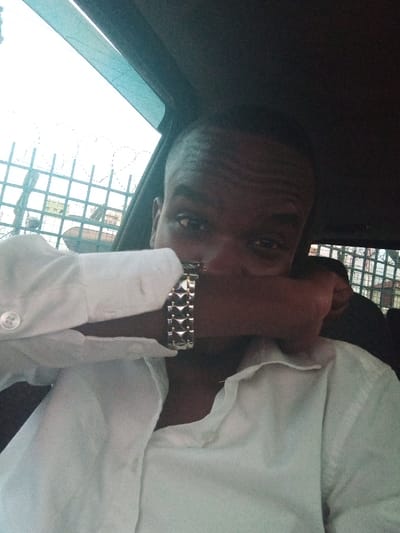 The image displays a dark-skinned male subject seated within a vehicle, located in Majengo ward, Kenya. He wears a white collared shirt, unbuttoned at the neckline, and a silver-colored metallic watch with a patterned link band on his left wrist. His left forearm and hand obscure the lower portion of his face, specifically his nose and mouth, while his eyes are visible and directed towards the camera. The dark interior of the vehicle's ceiling and trim is visible above and to the right of the subject. Through the vehicle's left window, an exterior environment is visible, characterized by a dark metal fence topped with barbed wire, set against a bright, overcast sky. Faint structural elements are discernible beyond the fence line.