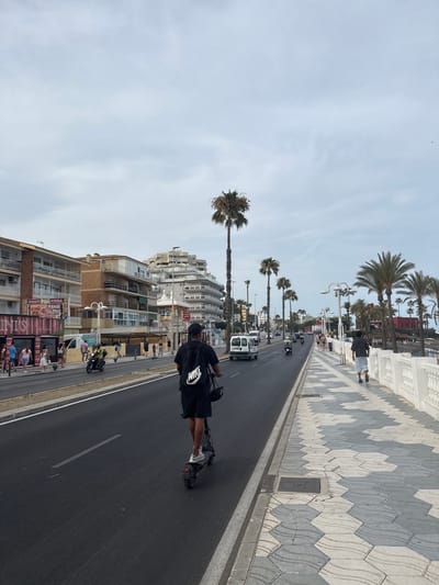 This media file depicts a street scene in Arroyo de la Miel-Benalmádena Costa, Spain, likely during the daytime under a cloudy sky. The camera is angled vertically, showing a road lined with palm trees and multi-story buildings.
The main subject in the foreground is a person riding an electric scooter. They are wearing a black t-shirt with a white Nike logo, black shorts, and white shoes. The scooter is also black and has a bag attached to the handlebars.
In the background, the road is a dark asphalt with white lane markings. Several other vehicles, including motorcycles and possibly a white van, are visible further down the road, suggesting moderate traffic. Pedestrians can be seen walking on the sidewalk next to the road, which is decorated with a distinctive hexagonal tile pattern in shades of gray and white. The buildings are a mix of modern apartment blocks and older structures, with balconies and windows characteristic of a coastal resort town. Some buildings have signs that are partially legible, with one appearing to advertise food. Palm trees are scattered throughout the scene, reinforcing the Mediterranean setting. The overall atmosphere appears to be a typical day in a busy tourist area.