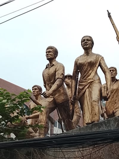 The image displays a large, multi-figure bronze-colored sculpture, identified as the "Shadhinota Shongram" (Freedom Struggle) monument, located on the campus of the University of Dhaka, Dhaka, Bangladesh. The sculpture depicts a dynamic group of individuals, predominantly men and women, cast in a matte, aged bronze-like finish, striding forward with determined expressions.

In the foreground, slightly left of center, a male figure in a collared shirt and trousers is depicted in mid-stride, looking upwards and to his left. To his immediate right, a female figure dressed in a sari holds a long rifle-like object vertically in her right hand and a rectangular object, possibly a book, in her left, also looking forward with a resolute gaze. Behind and around these central figures are additional statues, some carrying similar weapon-like objects, contributing to the impression of a unified march or struggle.

The base of the sculpture, made of concrete or stone, is partially visible. Multiple black electrical or communication wires are strung horizontally across the lower mid-ground, slightly obscuring the bottom portion of the monument. A cluster of green foliage from a tree or bush is visible on the left side, partially in front of some of the figures. In the background, a brown-tiled building with a sloping roof is discernible on the upper left, while the majority of the upper frame consists of a light, overcast sky. A vertical pole-like structure extends upwards from the top right edge of the frame. The perspective is a low-angle shot, looking up at the monument.
