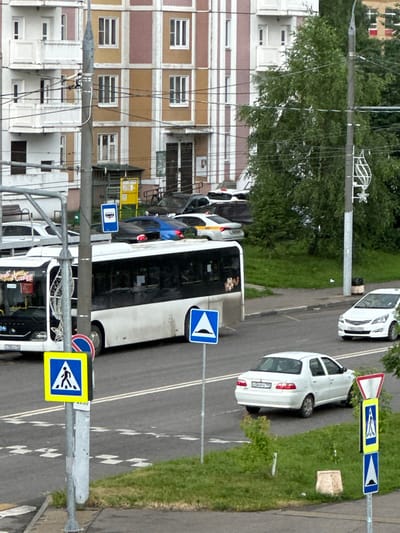 A wide-angle, eye-level outdoor shot captures a street scene in Podolsk, Russia. The foreground is dominated by a silver sedan driving away from the viewer, with a pedestrian crossing signpost to its left. The signpost features a yellow square with a pedestrian symbol and a blue square with a white upward-pointing triangle. Behind the sedan, a white bus is visible, its front on the left side of the frame. The bus has writing on its windshield and side. Further back, a blue car is parked. The background includes a multi-story building with a facade of beige and pink sections, several windows, and balconies. Trees with green foliage are present to the right of the building. The street is paved, with white lane markings visible. The sky is overcast.