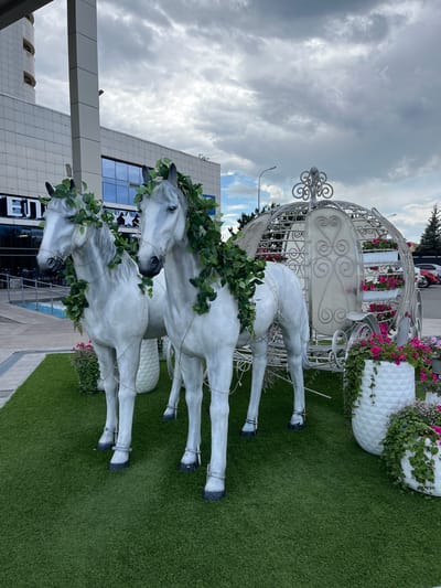 The image shows a decorative display featuring two white horse statues harnessed to a white carriage. The statues and carriage are adorned with green foliage and string lights. The display is set on an artificial green turf surface, with potted plants containing pink and white flowers placed around it. In the background, a modern building with a glass facade and a rounded architectural element is visible. The sky is overcast with gray clouds. The location is identified as being in the city of Kazan, Russia.