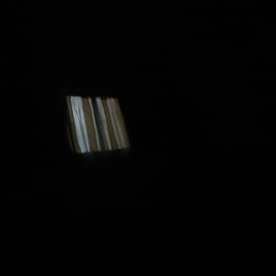 A close-up, low-light photograph shows a section of a striped curtain in what appears to be a dark room. The curtain exhibits alternating vertical stripes of light gray and a yellowish-brown hue. The lighting suggests it may be nighttime or a dimly lit interior space. The image is primarily black, with the curtain occupying a small portion of the frame in the lower-left quadrant. No other objects, people, or environmental context are visible. The location in the city of Zvertiv, Ukraine, cannot be determined from the image content.