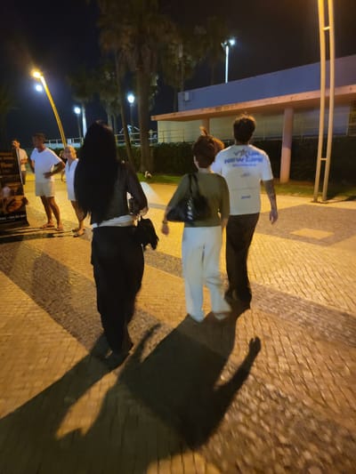 At night, on a cobblestone sidewalk in Quarteira, Portugal, near the marina, a group of four individuals walks away from the camera. In the foreground, a woman with long, dark hair wears a black sheer top and a long black skirt, carrying a black bag over her shoulder. To her right, a man in a white t-shirt with blue lettering and dark pants walks. To the left of the woman in black, a woman with short brown hair wears a green shirt and white pants, carrying a black bag. Further in the background, two men and a woman are visible near a lamppost and palm trees. One man wears a white t-shirt and shorts, while another wears a white t-shirt. A woman in a white top stands between them. The scene is illuminated by streetlights and the ambient light from nearby buildings.