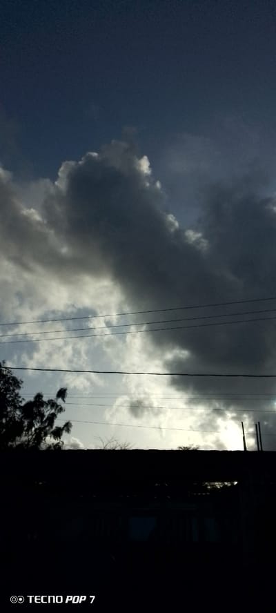 The image is a vertically oriented, dark photograph of the sky and a silhouetted foreground. In the upper portion of the frame, dramatic, heavy, dark grey clouds dominate the sky, with lighter grey and white clouds visible through breaks. A few thin, black power lines are strung horizontally across the middle of the image. The bottom third of the image is a silhouette of a dark structure, possibly a building or roofline, with some sparse, leafless tree branches visible against the sky above it. A bright light source, likely the sun, is peeking through the clouds near the right edge of the frame, casting a glow behind the silhouettes. The bottom left corner of the image contains the text "TECNO POP 7". The location in Altagracia, Venezuela, is not discernible from the visual elements in the photograph.