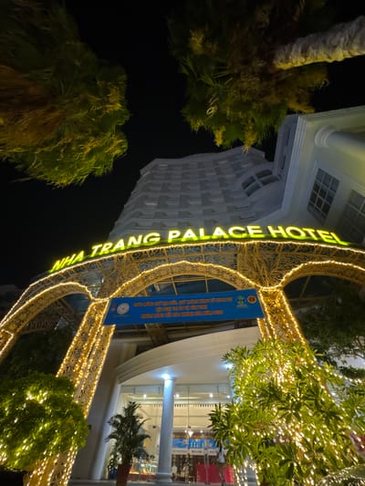 This media file depicts the exterior of the Nha Trang Palace Hotel at night, adorned with festive yellow lights. The hotel building is tall and modern, with a white facade and prominent columns. Palm trees with lush green fronds frame the entrance, and decorative lights trace the architectural details of an archway leading into the hotel. A blue banner hangs from the archway, displaying text in Vietnamese, including "CHÀO MỪNG QUÝ ĐẠI BIỂU, QUÝ KHÁCH HÀNG VỀ THAM DỰ" and "KHÁCH HÀNG YẾN SÀO KHÁNH HÒA NĂM 2026," suggesting an event is being hosted. The overall scene conveys a welcoming and celebratory atmosphere, illuminated by the warm glow of the lights against the dark night sky.