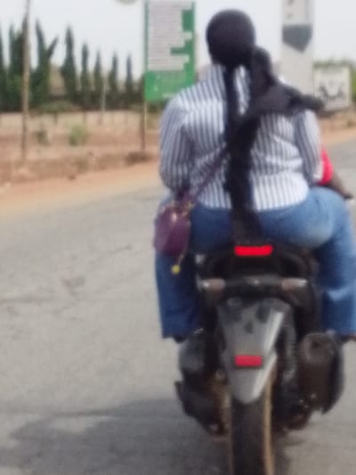 The image presents a rear view of a person riding as a passenger on a motorcycle on a road in Dungu, Ghana. The passenger is wearing a light-colored, vertically striped shirt (white and possibly light blue or grey), blue jeans, and a dark head covering with long, dark hair or fabric cascading down their back. A small, reddish-purple shoulder bag is visible hanging from their left side.

The motorcycle itself is dark-colored, seen from the rear, and features a prominent red taillight. What appear to be side bags or panniers are attached to the rear of the vehicle. The road surface is grey, possibly asphalt, and stretches into the background.

The setting is outdoors during daylight hours, under what appears to be an overcast or hazy sky. In the background, a line of tall, narrow trees or bushes is visible, along with several blurry signposts or utility poles, some showing hints of green signage. The overall image is somewhat blurry, which could be due to motion or a shallow depth of field. No visible text is legible on any signs, clothing, or the motorcycle itself.