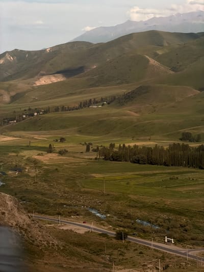 This is an aerial shot of a mountainous, rural landscape in Karagai Bulak, Kyrgyzstan. The scene is dominated by rolling green hills and valleys, with a winding road cutting through the terrain. A small, blue-roofed building is visible near the road, along with a red car parked on its side. Further down the road, a white structure resembling a house with a brown roof is also present. A narrow, rocky stream flows alongside the road in some sections. The hills are covered in sparse vegetation, with a line of trees marking a distinct boundary on one of the slopes. The sky is overcast, suggesting a cool, possibly windy day, with diffused light. The overall impression is one of a remote and natural setting, likely captured during daylight hours. There is no visible text in the image.