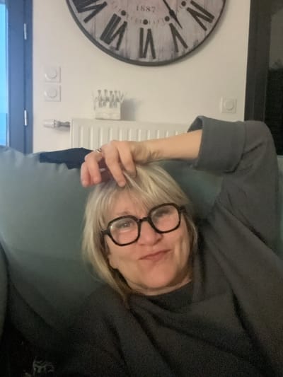The image depicts a woman with blonde hair and glasses lying on a gray or green surface. She is wearing a dark gray top. Her right hand is near her face, appearing to touch her hair. In the background, there is a wall with a large, circular clock displaying Roman numerals and the year 1887. Also visible are a white radiator, electrical outlets, and a small decorative item on the wall. The location, Chambœuf, France, is not verifiable from the image content alone.