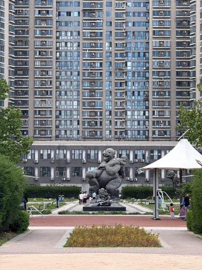 This image shows a section of the Changping District, China, focusing on its urban landscape and public spaces. The prominent features are two large buildings with a repetitive pattern of windows and air conditioning units, indicating a high-density residential or commercial area.

In the foreground, a public park or plaza is visible, equipped with exercise machines and a stone sculpture. There are a few people, including children, using the space, suggesting it is a recreational area for local residents. The scene is captured with a slightly unusual perspective, possibly due to the camera angle or rotation.