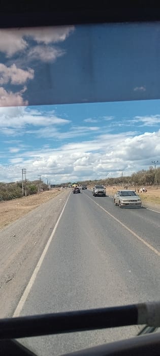 Captured from within a vehicle, a highway stretches into the distance. Several cars are spaced apart on the road. Fluffy white clouds fill the blue sky along the left side of the shot. Power poles run parallel to the road on the left. Some roadside vegetation can be seen along the edge of the asphalt. The shot is somewhat askew. A dark-colored frame is visible on the left and right sides.
