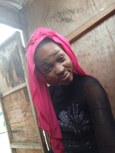 Adankolo, Nigeria: A young woman with dark skin is depicted in a close-up, slightly tilted portrait, smiling faintly. She wears a bright pink fabric head covering, loosely draped, and a black, long-sleeved sheer top featuring a prominent silver-toned, fan-shaped embellishment on the chest. The background consists of unpainted, weathered wooden planks, suggesting a simple interior structure. An opening, possibly a doorway or window, is visible on the left, leading to a brighter exterior. Faint, light-colored graffiti or markings, including alphanumeric characters such as "P804H2I3 F0", are visible on the upper right wooden panel, with a horizontal wooden beam below it.