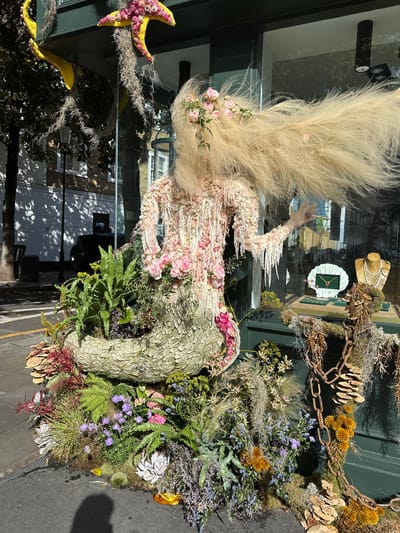 A window display in London, United Kingdom shows an elaborate floral arrangement and mannequin inside of a glass window. The mannequin is adorned with feathers and pink floral embellishments, with its arm raised. Adjacent to the mannequin is a large, round floral arrangement featuring ferns, moss, and various purple, pink, and green flowers. A chain is placed atop the arrangement. A yellow starfish is attached to the upper left corner of the window. Reflected in the window are building facades and trees. Inside the window, a display of jewelry is visible, with a mannequin wearing a pearl necklace and a white oyster shell with a green necklace.