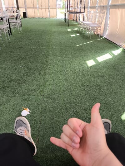 The image shows a person's right hand making a "hang loose" gesture on a green artificial turf surface. The person is wearing black pants and white and grey sneakers. There are several white chairs and a table in the background. The location is in Tatyr, Kyrgyzstan, based on the coordinates (latitude: 42.678, longitude: 74.656). The environment appears to be a covered outdoor space. The person is the only visible subject, and the environment suggests a relaxed, potentially recreational setting.
