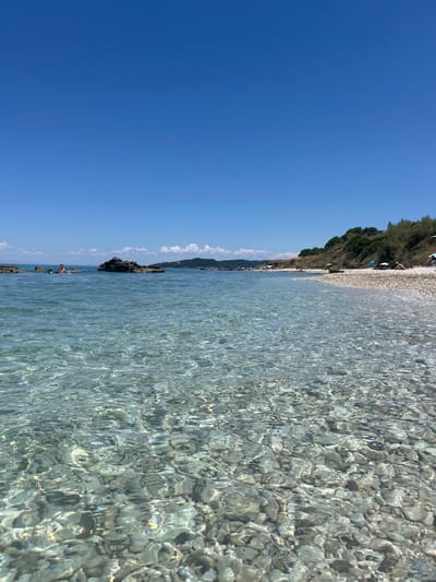 The image depicts a sunny day at a pebble beach in the Elaties Municipal Unit, Greece. Crystal-clear shallow water with a visible pebble seabed dominates the foreground. In the mid-ground, a few individuals are present in the water and on the shore. To the left, two people are partially submerged in the water, with one appearing to be wearing a snorkeling mask. Several rock formations emerge from the water. The beach itself is comprised of smooth, light-colored pebbles. Beyond the beach, a grassy, tree-covered hillside rises, with scattered white umbrellas and more people sunbathing. The sky is a clear, vibrant blue, indicating a cloudless day. The overall scene conveys a peaceful and inviting atmosphere of a seaside vacation.