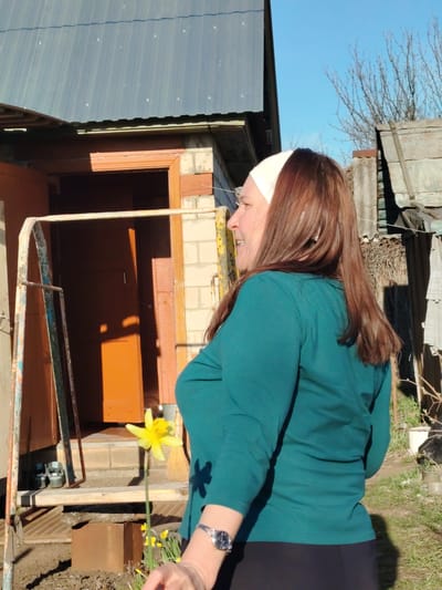 The image depicts a woman standing outdoors in what appears to be a backyard setting. She is wearing a turquoise long-sleeved shirt, black pants, and a white headband. She has reddish-brown hair and is wearing a wristwatch. She is facing to the left, smiling, and her arm is extended. In the foreground, there is a yellow daffodil flower. Behind her is a small building with a blue corrugated metal roof and an orange door. There is also a metal frame structure near the building. The environment includes grass, trees without leaves, and other outbuildings. The scene suggests it is a sunny day. The location is identified as Sarapul, Russia, based on prior context or knowledge.