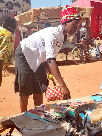 The image depicts a woman, wearing a white shirt and black skirt, holding a red and white checkered purse, and a small yellow fruit, standing near a table covered in assorted combs and hairbrushes. The woman is looking down at the table, possibly selecting a comb or brush to purchase. Behind the table, a bicycle sits parked nearby, with a red building in the background. The scene takes place in a marketplace in Lwandeti, Kenya, near the location with coordinates (latitude: 0.563, longitude: 34.806). The setting appears to be a bustling, open-air market with various vendors selling their wares. The woman is the main subject of the image, while the table of combs and brushes and the bicycle are secondary subjects. The overall atmosphere of the scene is vibrant and active. The image appears to have been taken in the daytime, with sunlight illuminating the scene. The ground is a mix of dirt and sand, typical of a market setting in Lwandeti, Kenya. The overall scene captures the everyday hustle and bustle of life in Lwandeti, Kenya, with a focus on the woman's transaction at the market.