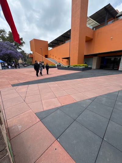 Here is a detailed description of the image:
**General Scene:** The image presents an outdoor urban setting, likely a plaza or pedestrian area in Mexico City, Mexico. The scene is captured from an elevated perspective, looking down at the plaza.
**Architecture:**
* The plaza is surrounded by modern, orange-toned buildings with strong geometric lines.
* The buildings have multiple levels, indicated by the presence of balconies and roof structures.
* There are visible windows and metal awnings, suggesting commercial or institutional purposes.
**Plaza Surface:**
* The plaza is paved with large square tiles of two contrasting colors: pinkish-red and dark gray, arranged in diagonal bands.
**People:**
* Several individuals are walking across the plaza. They are dressed in dark clothing, likely casual attire.
* The people appear to be engaged in normal pedestrian traffic, walking towards or away from the buildings.
**Vegetation:**
* There is a purple blooming Jacaranda tree visible, a characteristic feature of Mexico City.
* Hedges and other greenery are planted around the buildings.
**Other Objects:**
* A portion of a red flag or banner is visible in the lower left corner, potentially indicating an event or decoration.
**Environment:**
* The sky is overcast, with diffuse sunlight illuminating the scene.
* The overall atmosphere suggests a typical day in an urban environment.
**Location Clues:**
* The architectural style, combined with the presence of Jacaranda trees, suggests the location is in Mexico City.
**Actions/Interactions:**
* People are walking around on the plaza.
* The scene captures a moment of daily life in a city square.
In summary, the image depicts a modern plaza with pedestrians in Mexico City, characterized by its distinct architecture, paved surface, and the presence of iconic Jacaranda trees.
