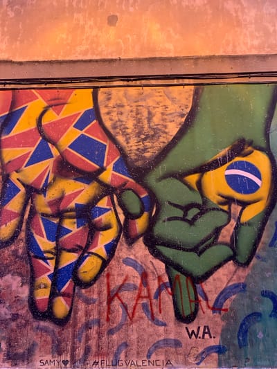 This image depicts a graffiti mural on a textured wall in Valencia, Spain. The mural features two hands clasped together, a symbol of connection or unity. One hand, depicted in green, holds a yellow orb emblazoned with the Brazilian flag. The other hand is a mosaic of geometric shapes in red, yellow, and blue, evoking a sense of vibrant energy. Below the hands, the graffiti artist's tag "SAMY" and a hashtag "#FLUGVALENCIA" are visible, along with the initials "W.A." The wall itself shows signs of wear and tear, with peeling paint and visible brickwork, suggesting an outdoor urban environment. The lighting suggests it is daytime, with a warm glow illuminating the upper portion of the wall.