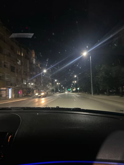 This is a nighttime, first-person perspective shot from inside a vehicle driving on a city street in Plovdiv, Bulgaria. The scene is illuminated by streetlights and vehicle headlights. On the left side of the street, there is a multi-story building with visible windows and some illuminated signs. The road ahead is wet, reflecting the lights. Several vehicles are visible on the road, including cars with their headlights on. Trees line the right side of the street. The dashboard of the vehicle is visible in the foreground.