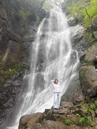 A medium, wide outdoor shot depicts a person posing in front of the Makhuntseti Waterfall in Kveda Makhuntseti, Georgia.
The waterfall cascades down a cliff face. The cliff is characterized by dark, rocky formations and patches of greenery.
A person is lying horizontally on a rocky ledge, appearing to pose in front of the waterfall. They are wearing a light-colored long-sleeved shirt and jeans. Their hands are positioned behind their head.
In the upper part of the frame, obscured by the angle and lighting, are hints of foliage and rocky outcroppings, along with parts of other people.