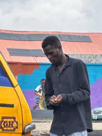 In Ajara, Nigeria, a young Black man is captured in a medium shot, looking down at his hands as he manipulates something small. He is wearing a dark, collared, long-sleeved shirt and blue jeans. The background features a colorful mural on a building with a red roof adorned with solar panels. To the left, the front corner of a yellow vehicle, possibly a tro-tro, is visible, displaying the text "GH 2000" and a Ghanaian license plate within an emblem. A mural depicting another person holding a basket is also present in the background, with a teal car parked to the far right. The sky is overcast.