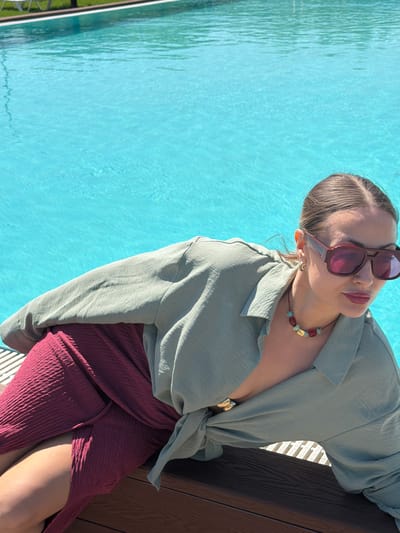 The image features a woman posing outdoors by a vibrant blue swimming pool on a bright, sunny day. She is shown from the chest up, leaning against a dark brown wooden surface, with her legs and a portion of her deep red skirt also visible. Her light brown hair is pulled back, and she wears large, reddish-brown framed sunglasses.
A colorful beaded necklace, featuring red, blue/turquoise, and gold-colored stones, is visible around her neck, complemented by a small gold-colored earring on her right ear. She is dressed in a light green, long-sleeved, collared shirt made of a textured, crinkled fabric, which is partially unbuttoned. Her deep red skirt, also of a textured, crinkled material, has a high slit.
The background is dominated by the clear, sun-dappled blue water of the swimming pool, exhibiting ripples and reflections. A metallic fence or railing is faintly visible on the far left edge of the pool. The bright daylight and clear water suggest a sunny weather and a relaxed, leisurely atmosphere, likely at a poolside location such as a resort or private residence. No text is visible in the image.